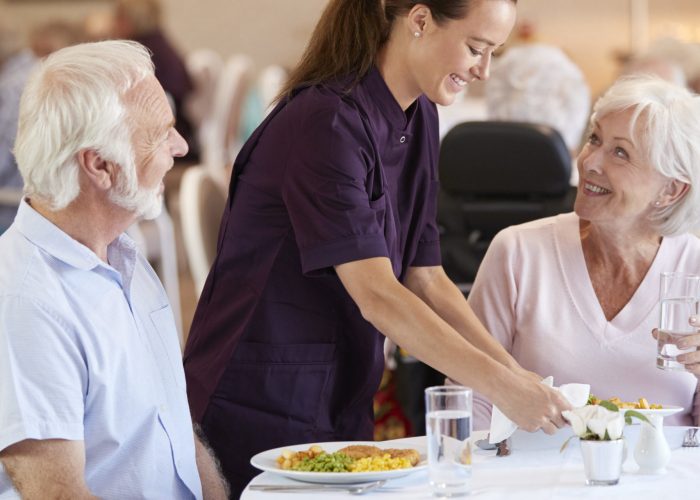 Senior Couple Being Served With Meal By Carer In Dining Room Of Retirement Home